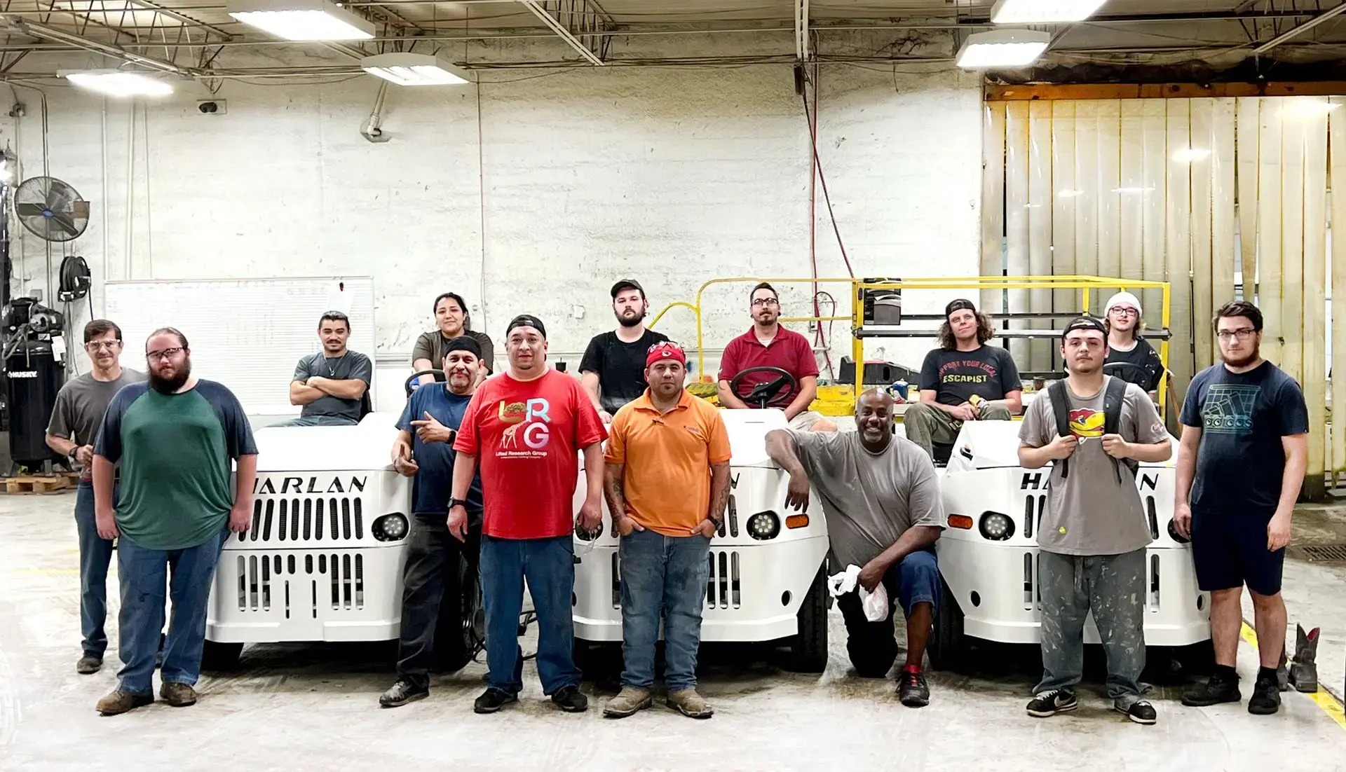 A group of Harlan mechanics in casual work attire poses together inside a brightly lit manufacturing facility