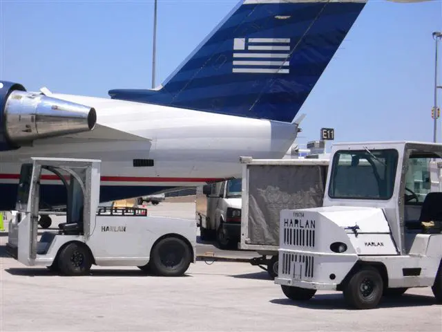 Two white Harlan tractors are parked on a sunny airport tarmac in front of the blue and white tail of a large commercial aircraft.