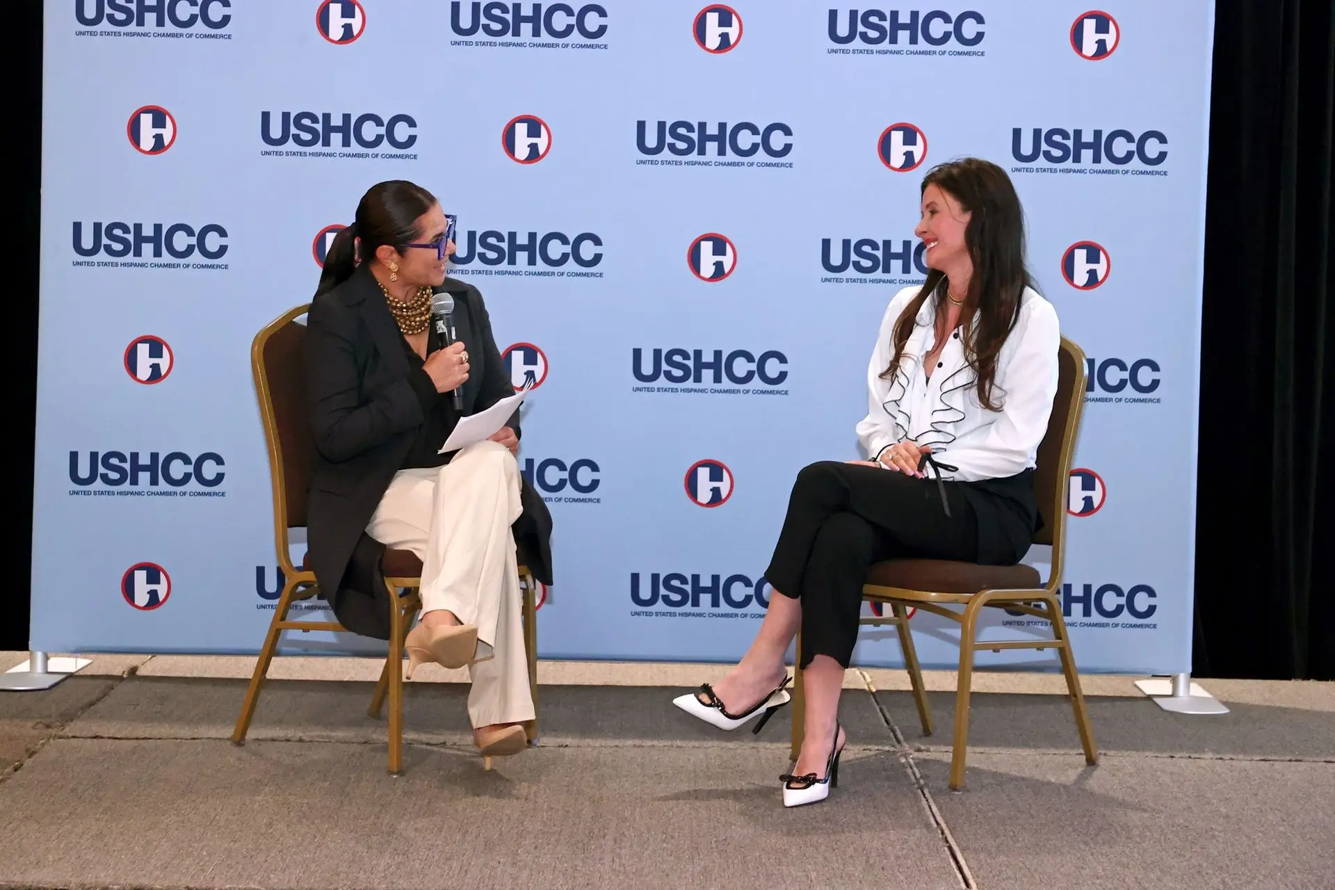 Two women sit in gold-framed chairs on a stage during an interview, positioned in front of a blue backdrop repeatedly branded with the USHCC (United States Hispanic Chamber of Commerce) logo.