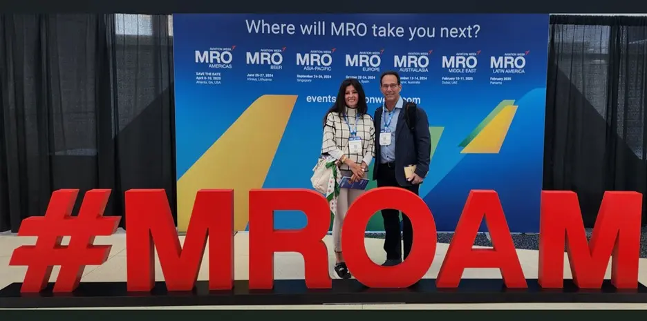 Harlan CEO Jamie Kaplan and a woman stand behind a large, 3D red floor sign that reads "#MROAM" in an indoor exhibition hall.