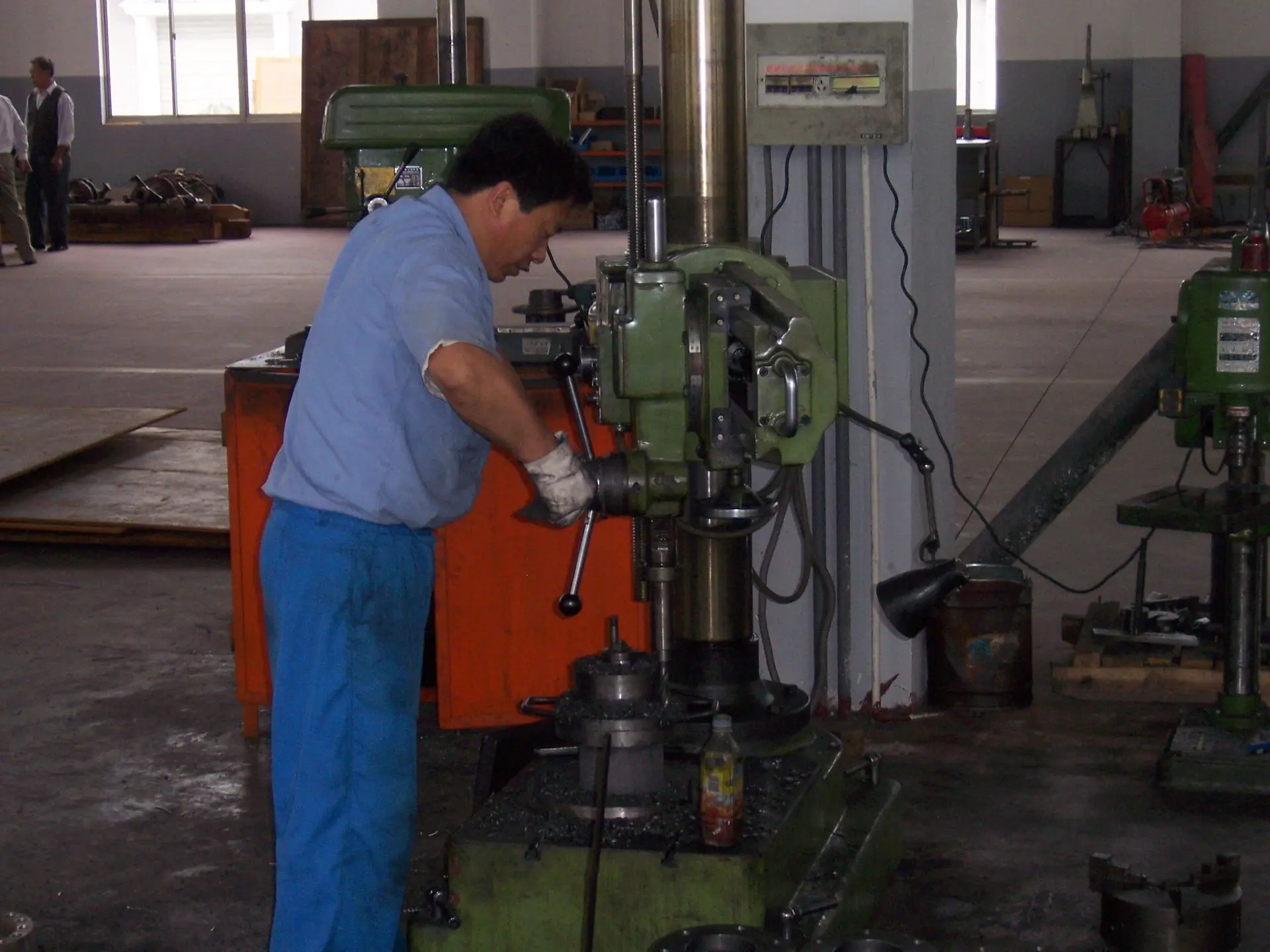 Industrial worker operating a heavy-duty radial drill press for manual metal hole boring in a machining workshop.