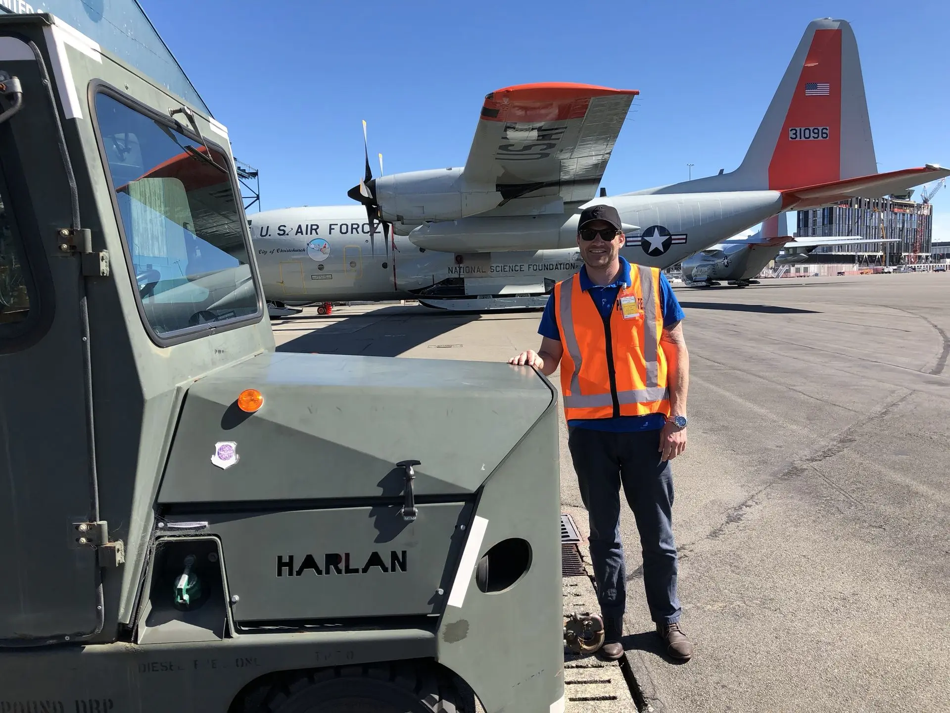 Ground crew member standing with a Harlan aircraft pushback tractor at an international airport, supporting a U.S. Air Force National Science Foundation flight for global polar research.